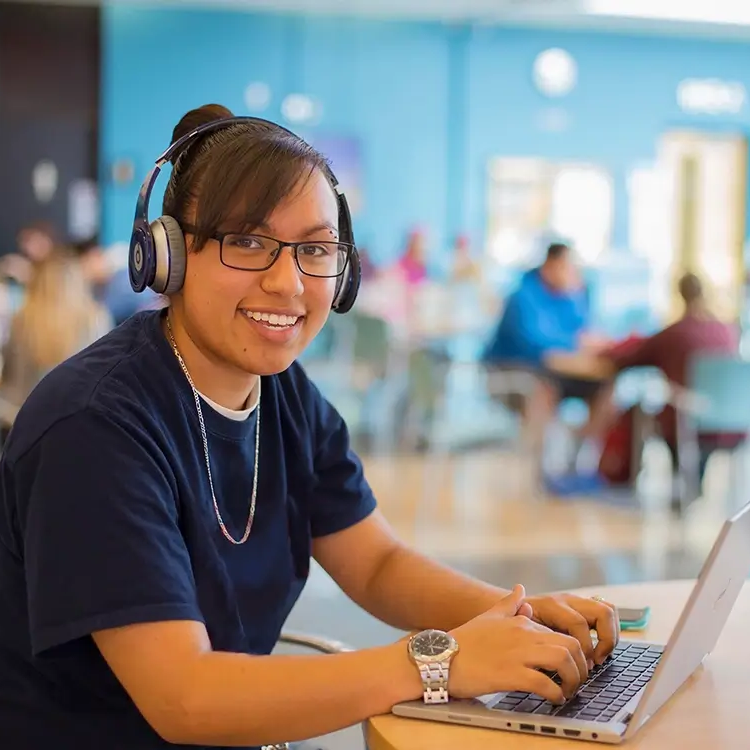 A student smiles at the camera sitting in a cafeteria on a laptop